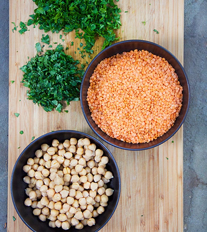 wooden cutting board topped with chopped herbs and two bowls, one filled with chickpeas the other with raw red lentils