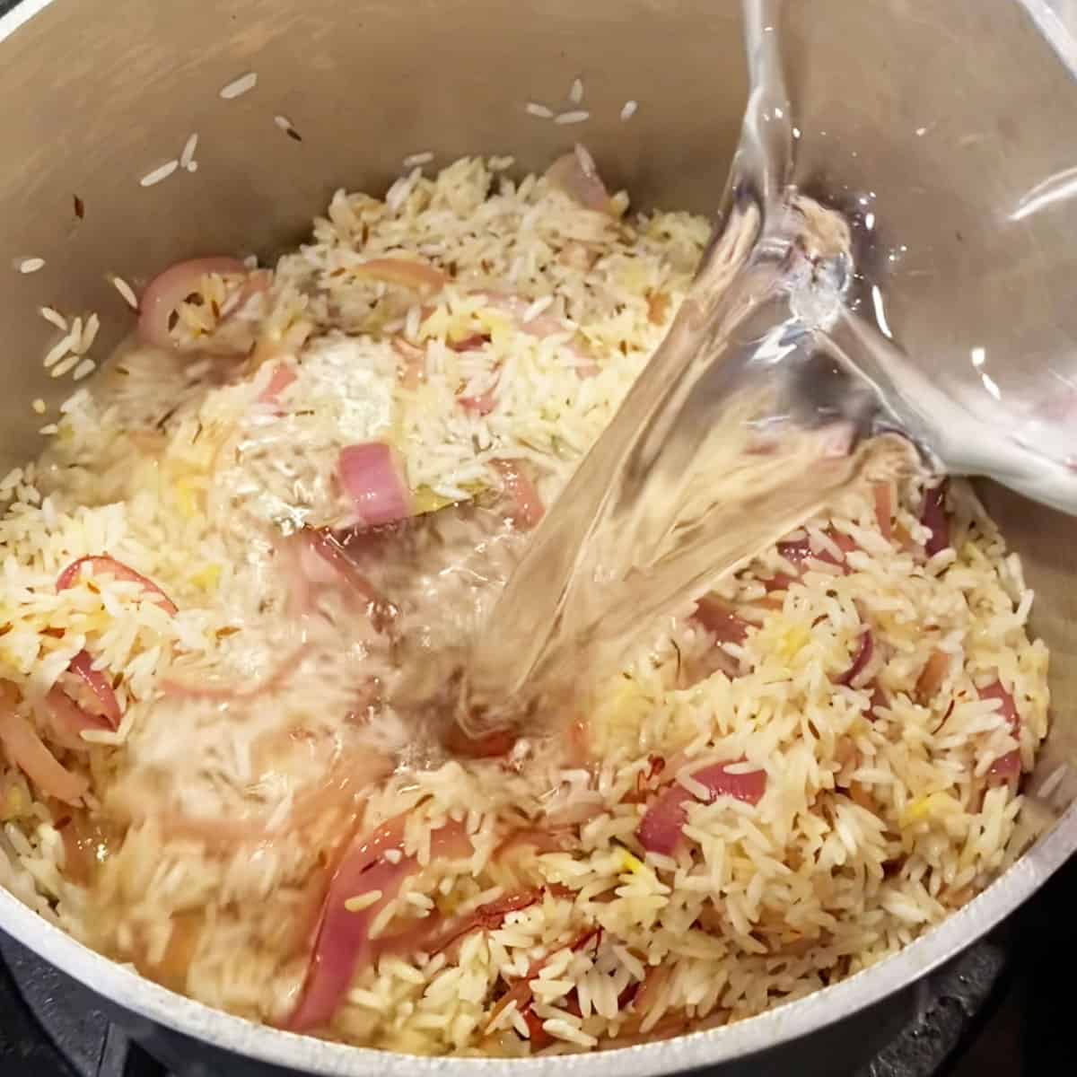 Small saucepan filled with rice, sauted nions and whole spices, water being poured in from a measuring cup.