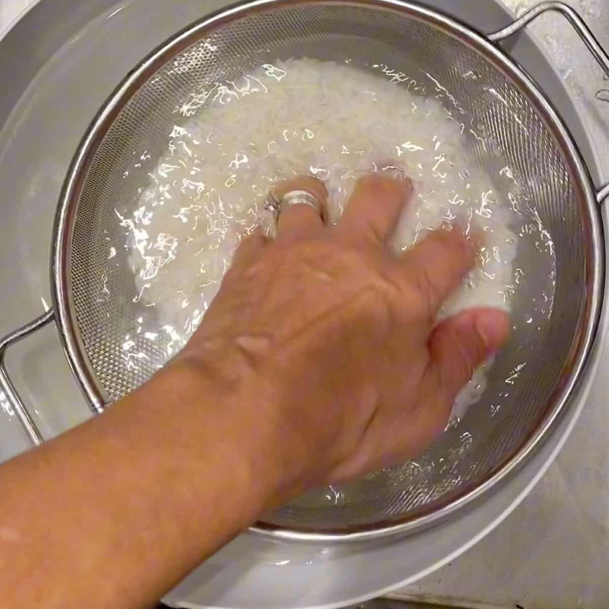 Rice in a colander filled with water, a hand swishing the rice around.