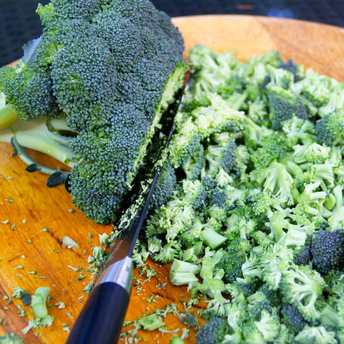 a head of broccoli being shaved with a large knife, into small pieces, on a wooden cutting board.