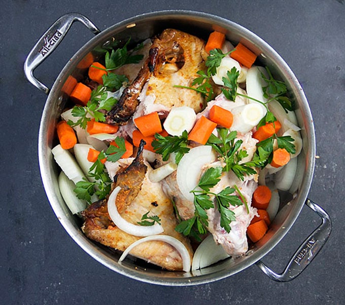 A soup pot seen from above, filled with ingredients to make turkey soup: hunks of leftover turkey carcass, chopped onions, carrots and parsley