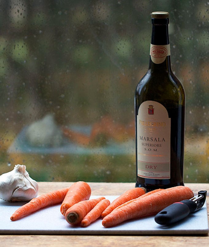 A half dozen carrots on a cutting board with a vegetable peeler and a bottle of Marsala wine in the background.