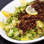 A white oval platter on a black background, filled with sautéed shredded Brussels sprouts topped with fried shallots, lemon slices decorate the rim of the platter