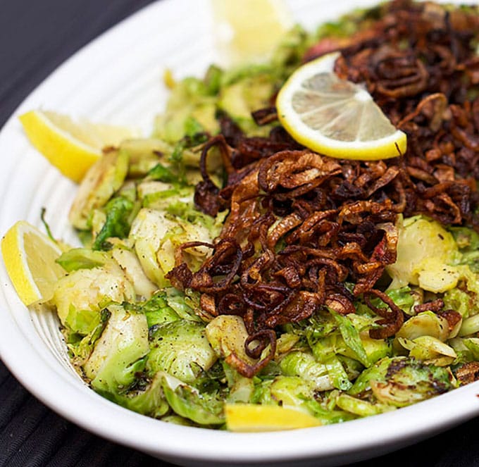  A white oval platter, on a black table cloth, filled with sautéed shredded Brussels sprouts topped with Fried Shallots, several slices of lemon decorating the rim of the platter.