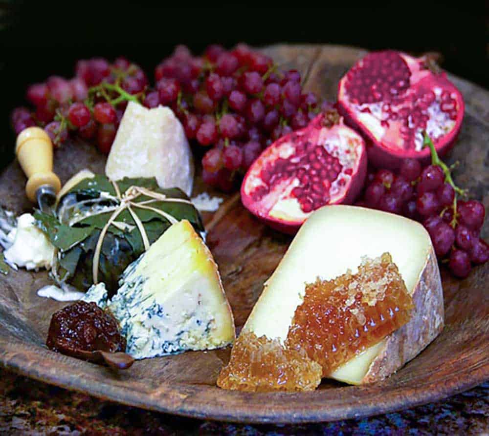 dark brown wooden cheese platter topped with cheeses, grapes, honeycomb and a halved pomegranate