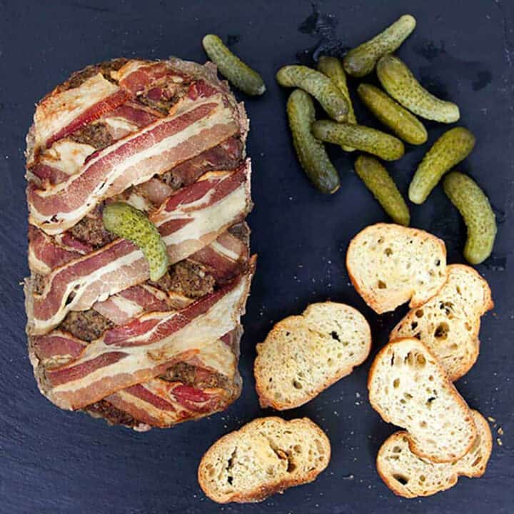 A loaf of pate wrapped with woven strips of bacon and a cornichon on top, with a handful of cornichons and a handful of crostini on the side.