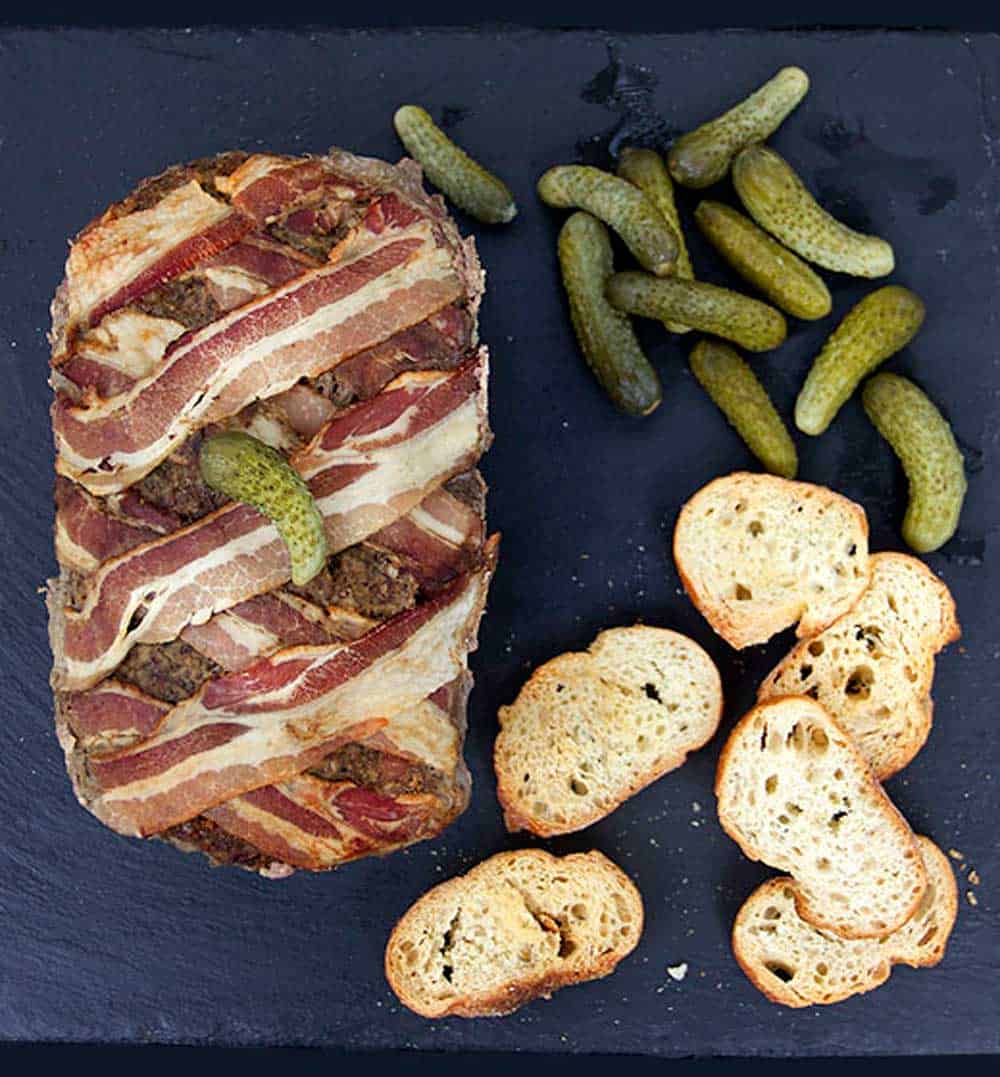 Homemade pate with a bacon lattice top next to a bunch of cornichons and sliced baguette