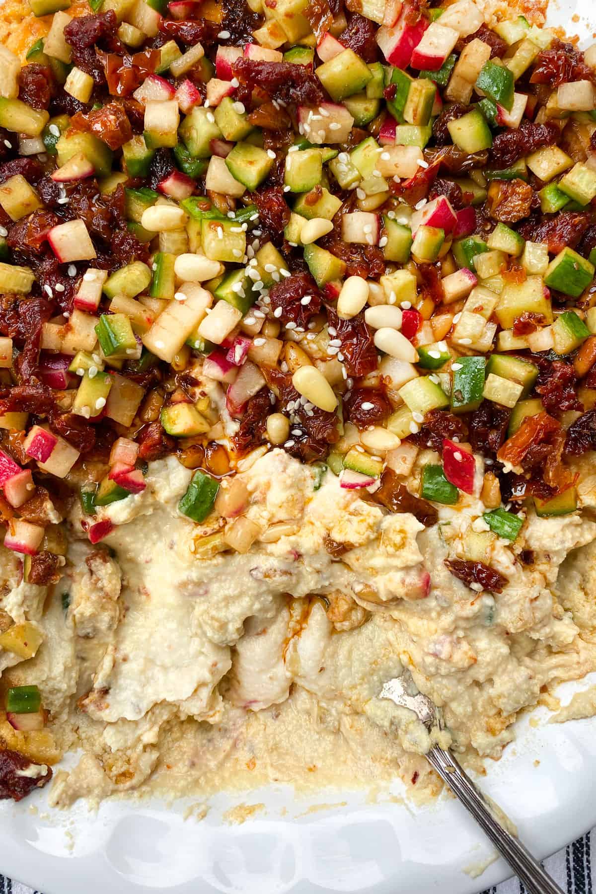 close up of creamy hummus topped with chopped cucumbers, radishes and sun dried tomatoes