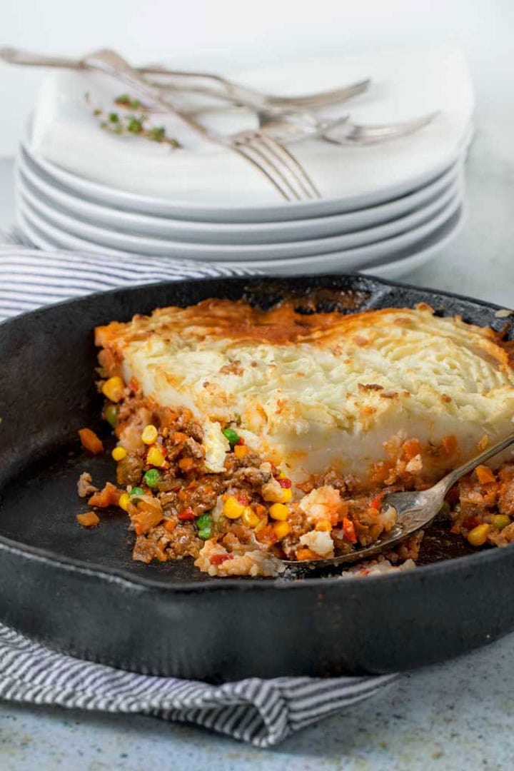 cast iron skillet with a large piece of shepherd's pie in it and a stack of plates and forks in the background