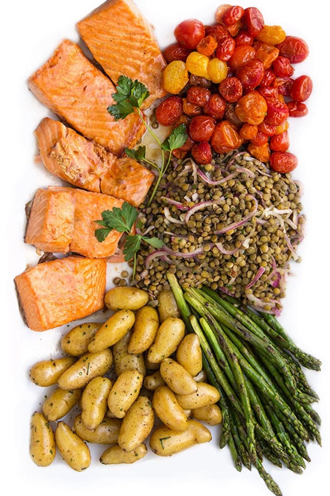 a composed salads on a white platter with five pieces of broiled salmon fillet, roasted cherry tomatoes, french lentils, sautéed thin asparagus and herbed fingerling potatoes