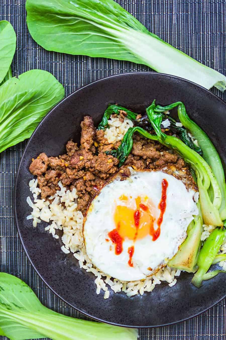 Thai Beef Rice Bowl in a shallow black bowl with stir-fried ground beef, brown rice, sautéed bok choy, and a fried egg on top. leaves of raw bok choy surround the plate.