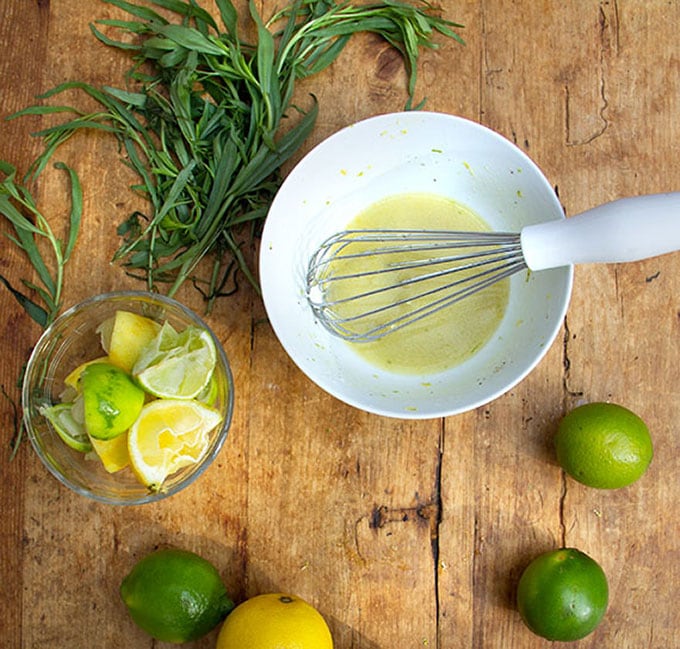 A whisk in a white bowl of citrus honey vinaigrette, surrounded by lemons and limes