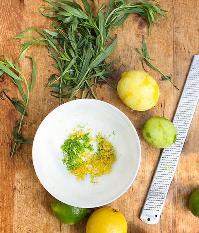 a zested lemon and a zested lime and a zester next to a white bowl of lemon and lime zest