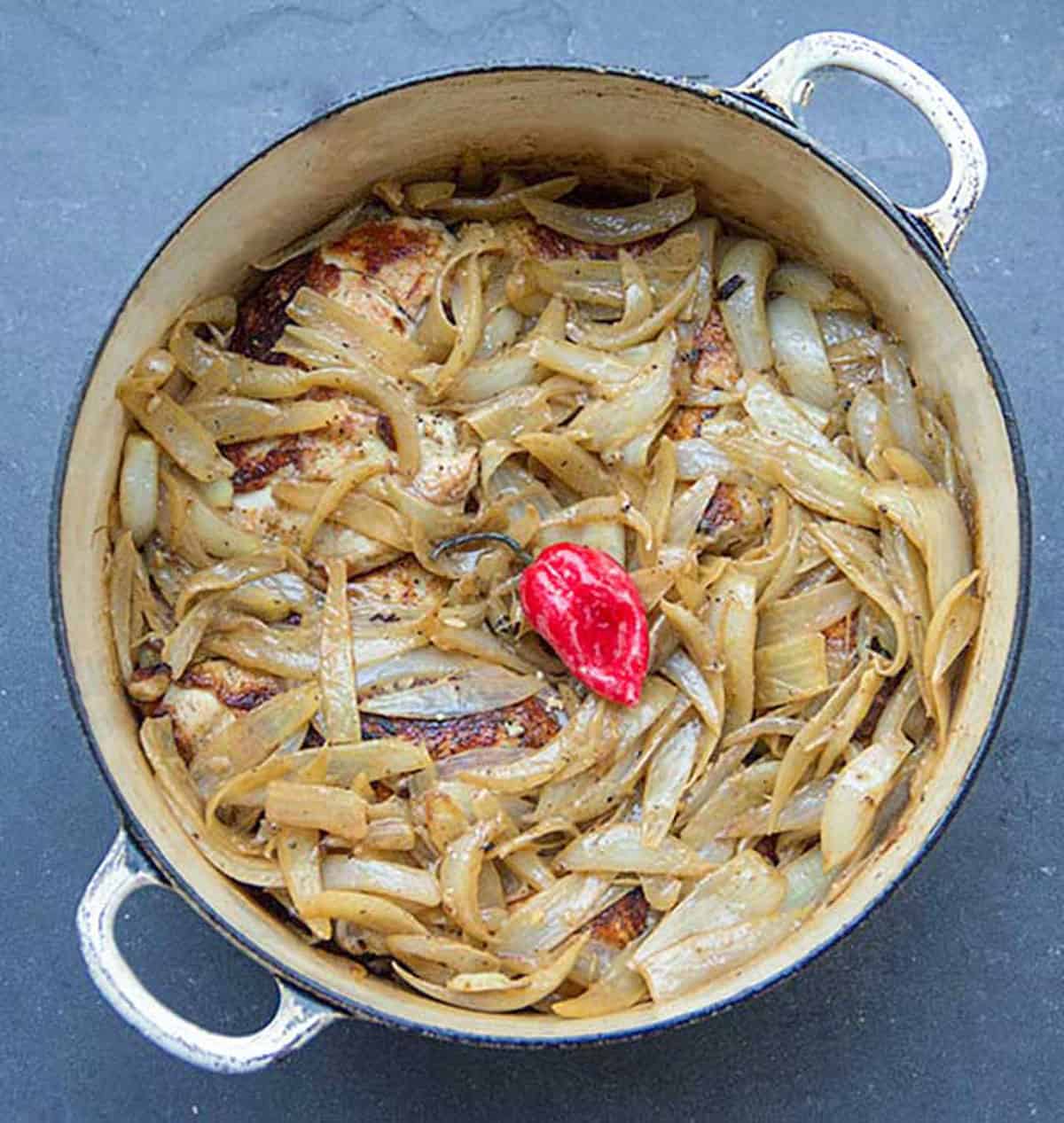 Dutch oven with a layer of caramelized onions sitting on top of browned chicken parts and a small spicy red pepper in the center.