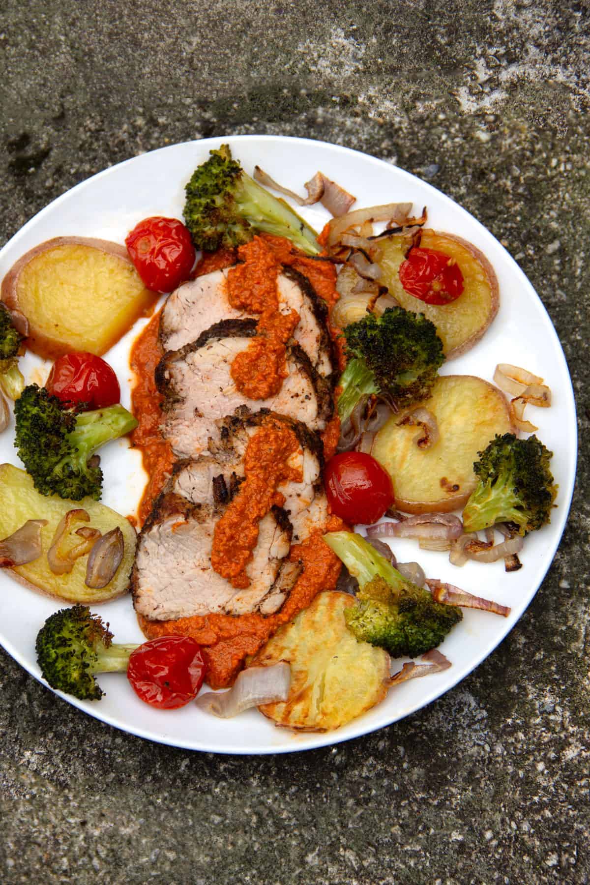 A plate with few slices of roasted pork tenderloin drizzled with Romesco sauce and surrounded by roused potaoes, broccoli and cherry tomatoes.