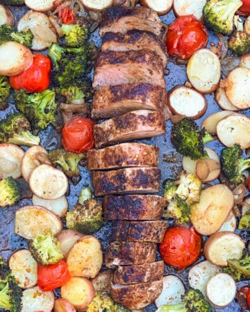 Sheet pan with sliced roasted pork tenderloin arranged down the center, surrounded by roasted potatoes, broccoli and cherry tomatoes.