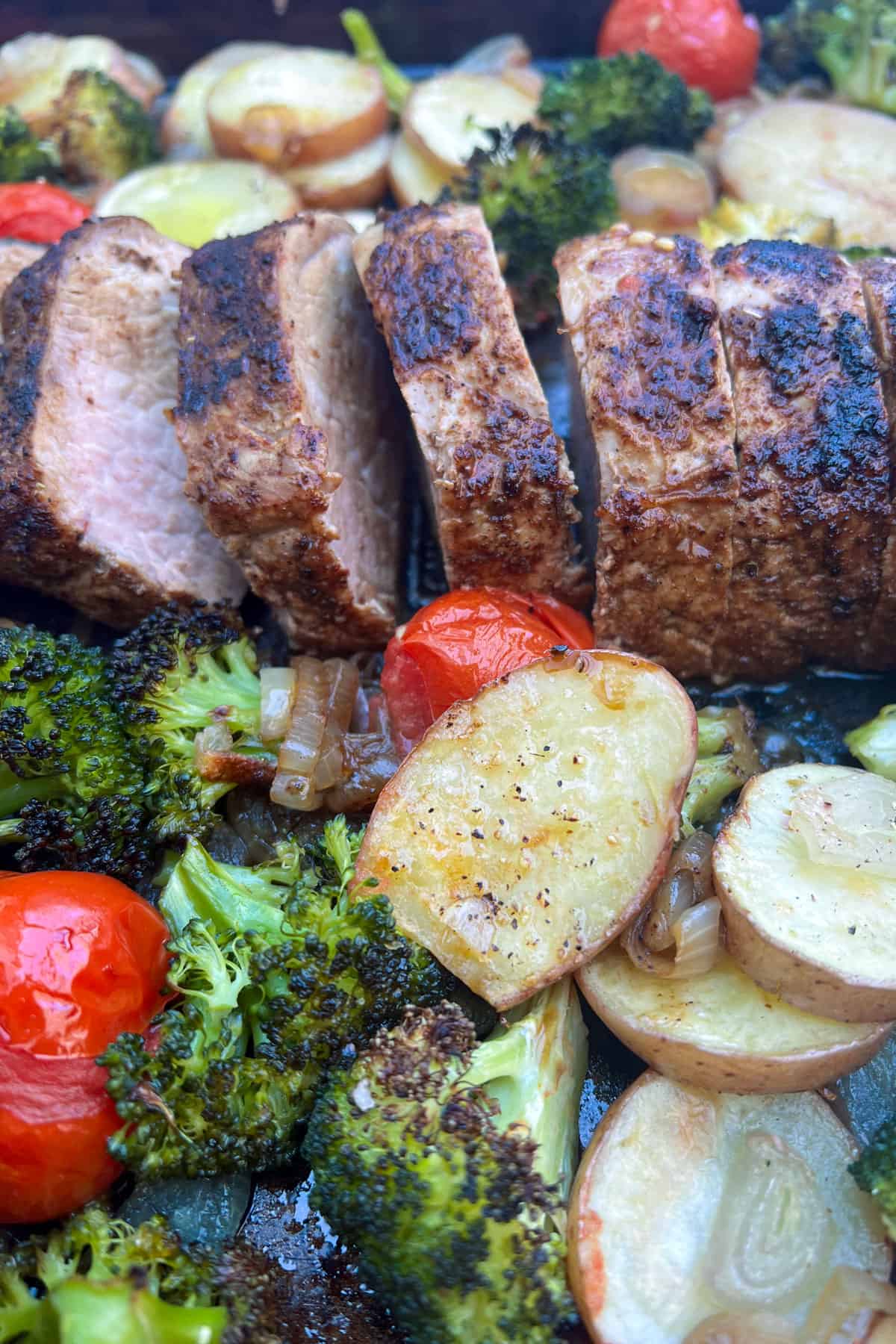 sliced, roasted pork tenderloin on a sheet pan with broccoli, sliced potatoes and roasted tomatoes.