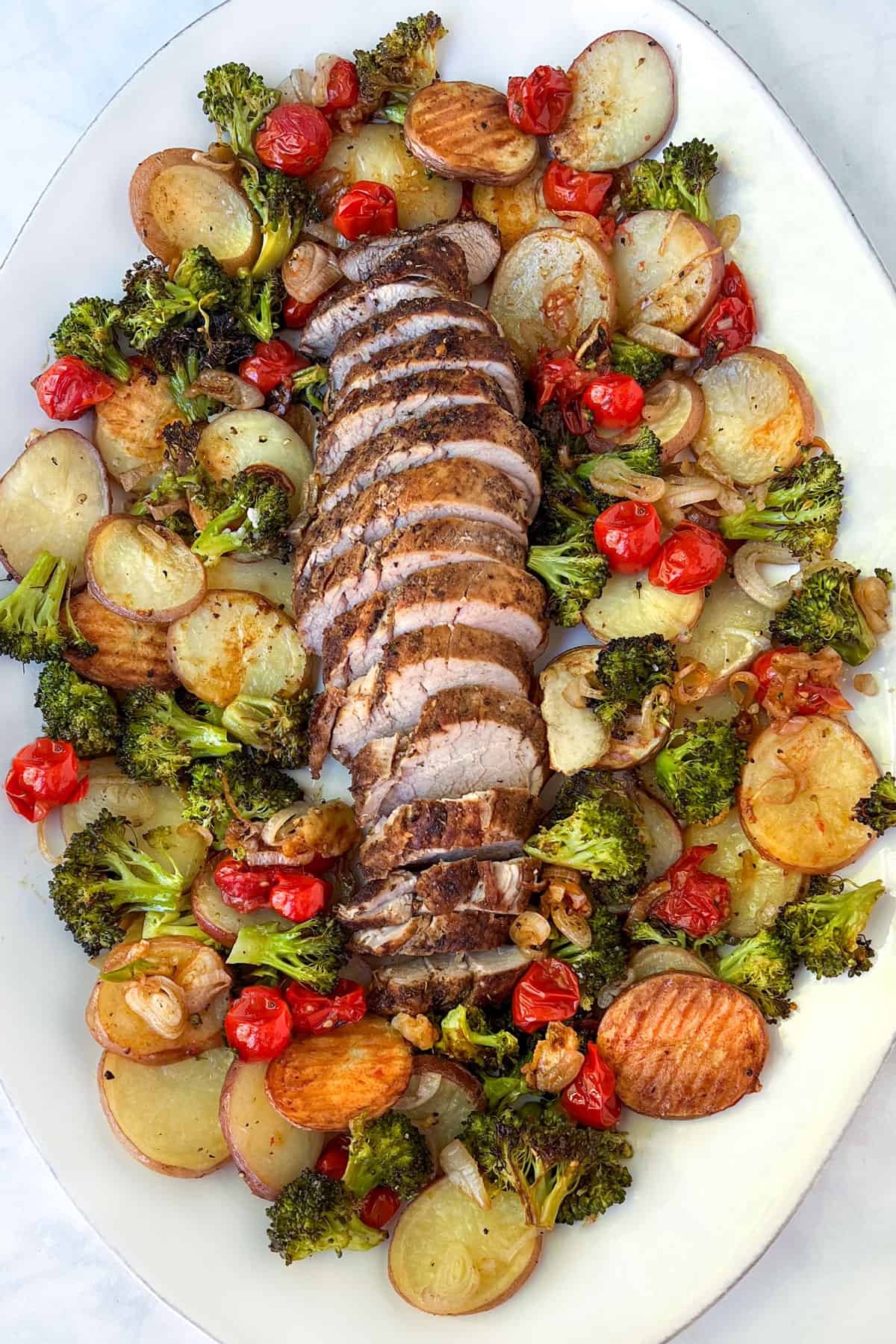 oval platter with sliced roasted pork tenderloin arranged down the center, surrounded by roasted potatoes, broccoli and cherry tomatoes.