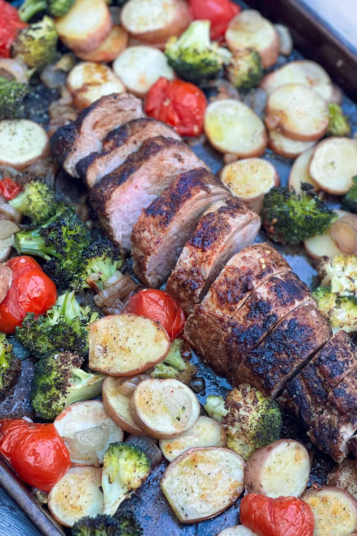 sliced roasted pork tenderloin on a sheet pan surrounded by sliced roasted potatoes, roasted cherry tomatoes and roasted broccoli