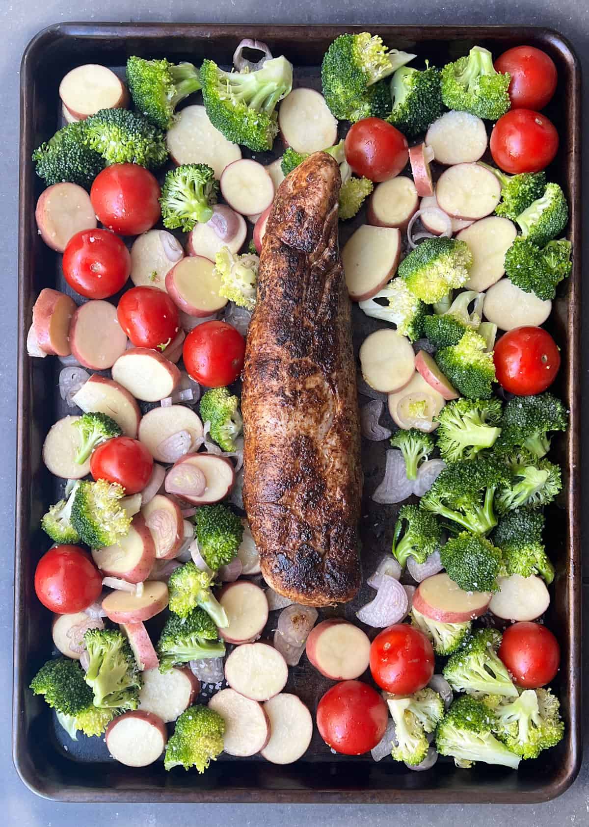 seared pork tenderloin on a sheet pan surrounded by raw broccoli florets, cherry tomatoes and sliced potatoes