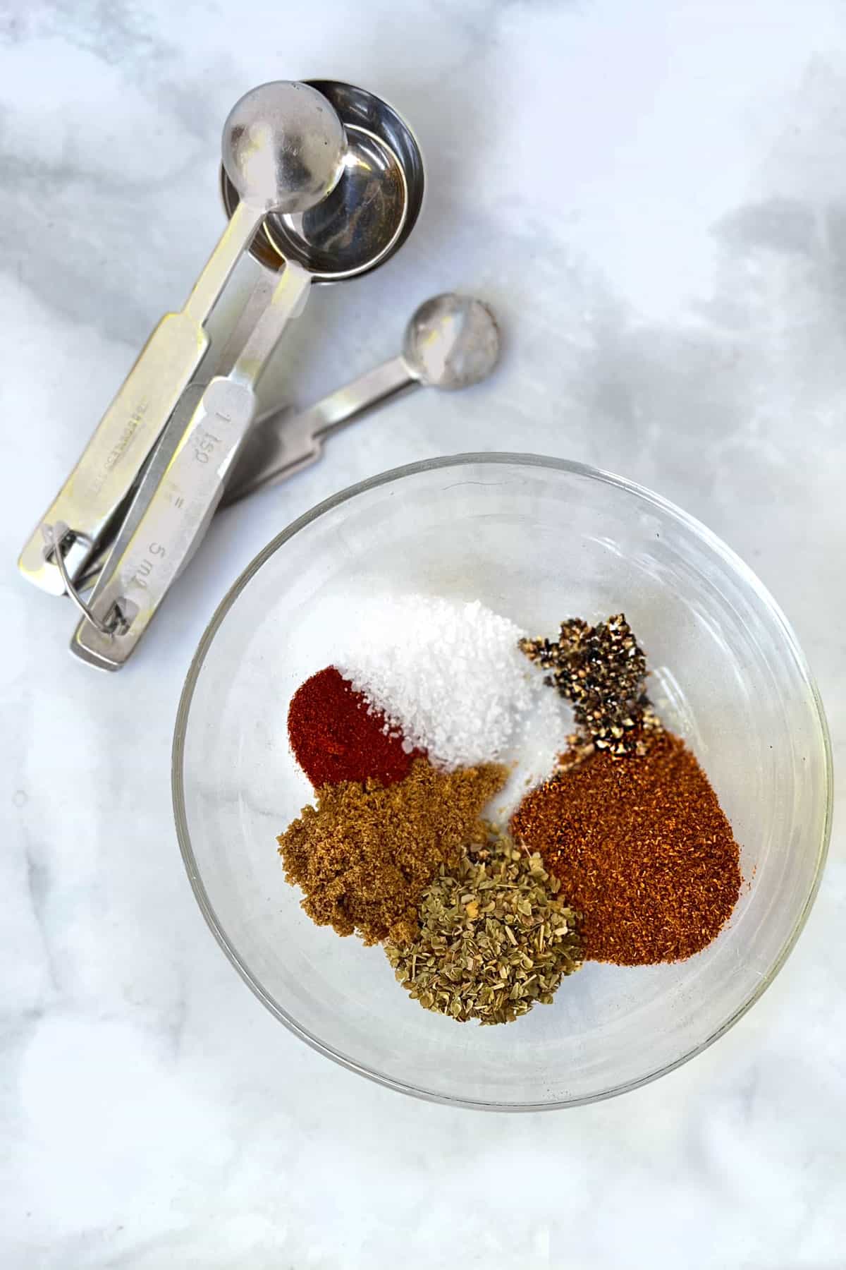6 different spices in a glass bowl next to measuring spoons