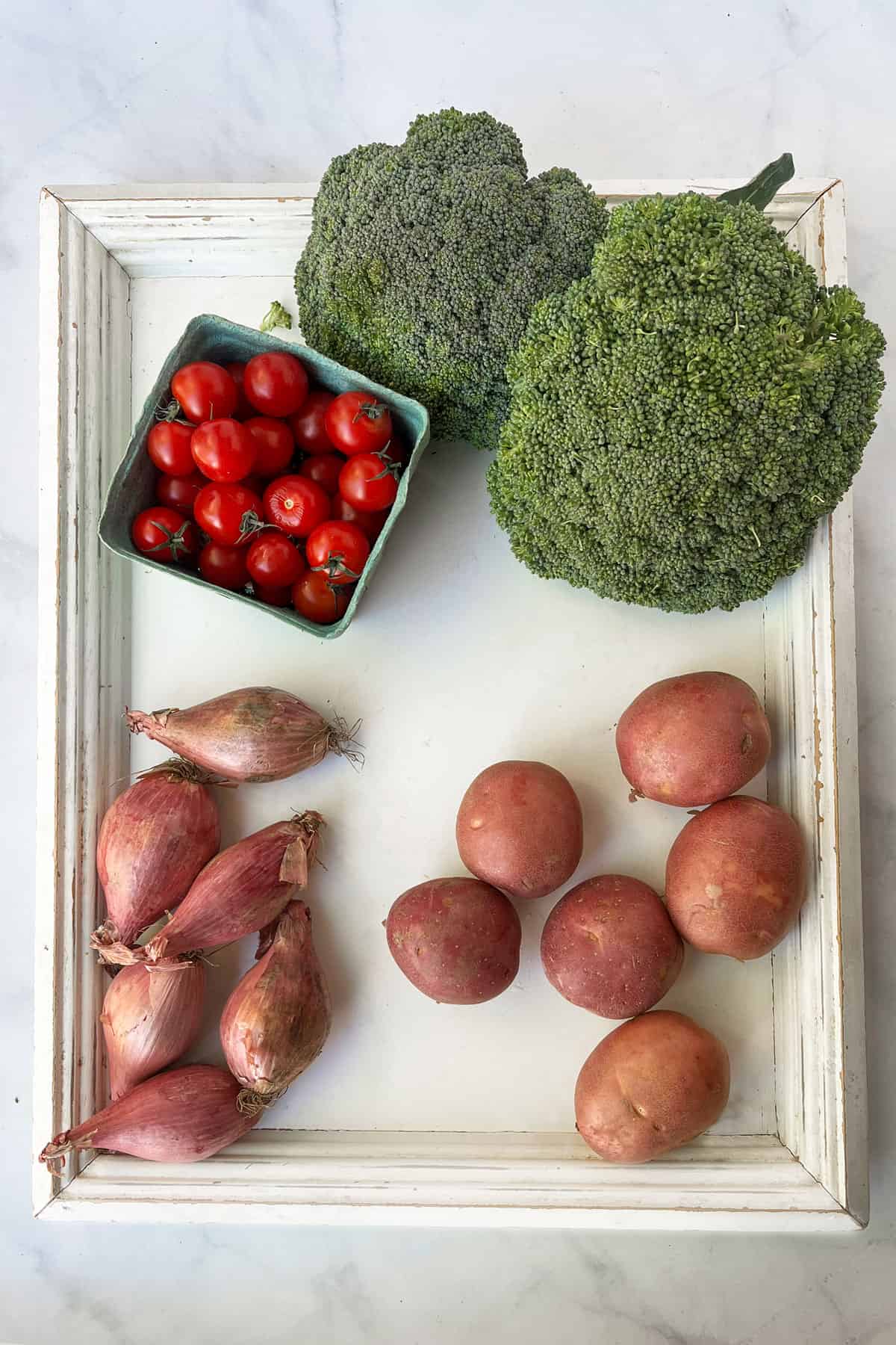 2 broccoli crowns, 6 red skinned potatoes, 6 shallots and a pint of cherry tomatoes on a white wooden tray