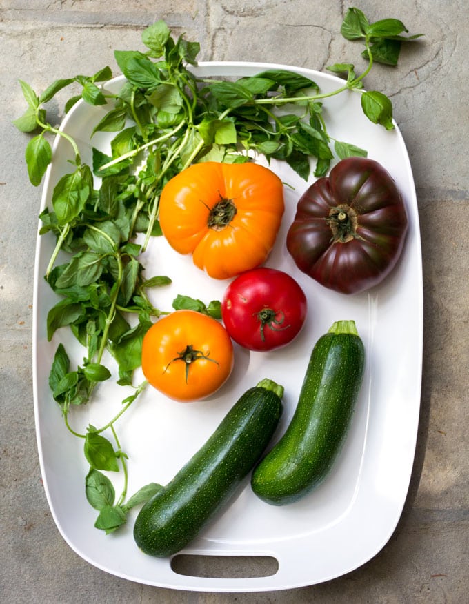 a white tray with ingredients for zucchini caprese stacks: two zucchinis, 4 heirloom tomatoes and a bunch of fresh basil