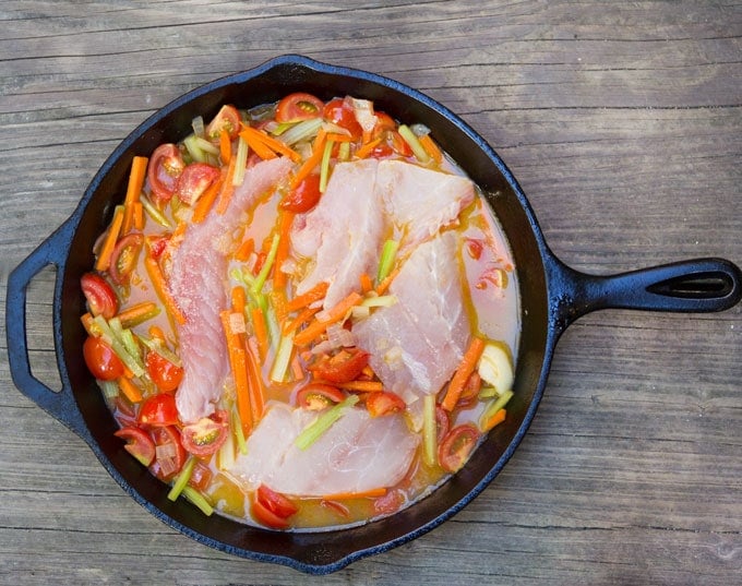 Cast iron skillet with matchstick cut carrots and celery, chopped onions, sliced cherry tomatoes, raw fish and water