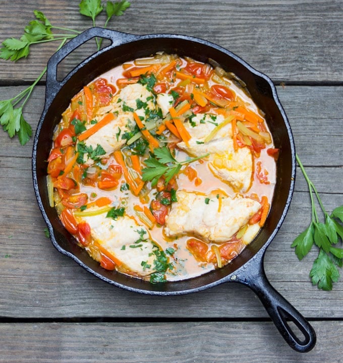 Cast iron skillet filled with fish, sliced vegetables, tomatoes and water. Garnished with parsley.