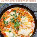 cast iron skillet filled with white fish fillets surrounded by chopped carrots and cherry tomatoes and broth and sprigs of parsley