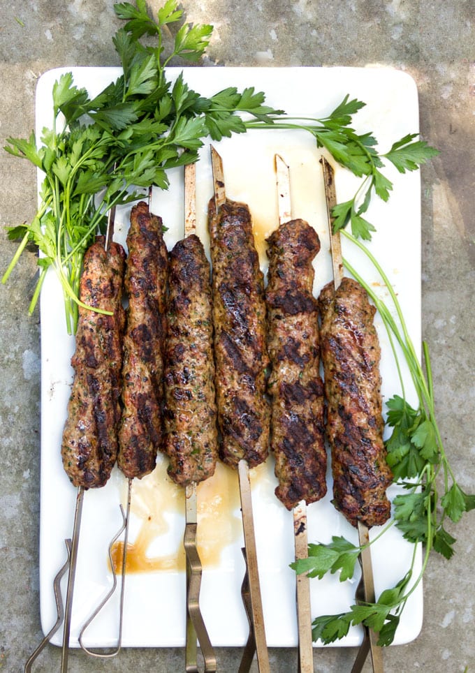 six lamb kofta kebabs on skewers on a white rectangular platter decorated with parsley