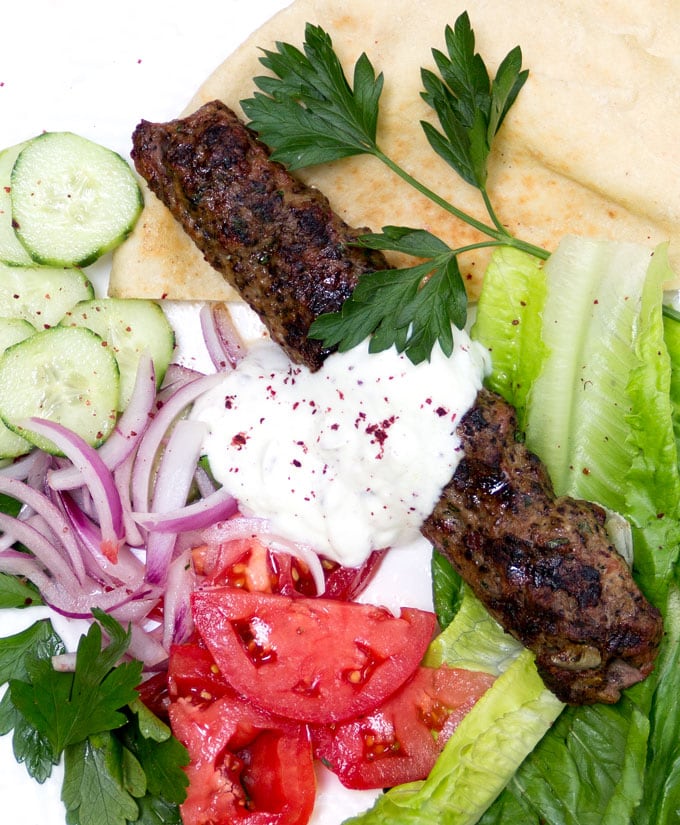 One grilled turkish Kofta Kebab on a pita with cucumber rounds, sliced red onion, sliced tomatoes, lettuce leaves, parsley sprigs, and a topping of yogurt sauce with sumac