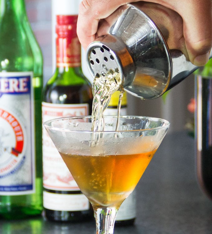 A metal cocktail shaker pouring a Perfect Manhattan into a martini glass