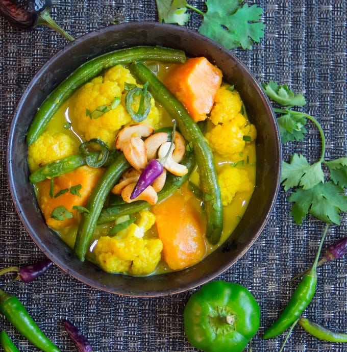 Black bowl surrounded with herbs and peppers and filled with green beans and chunks of cauliflower, sweet potato in yellow curry sauce