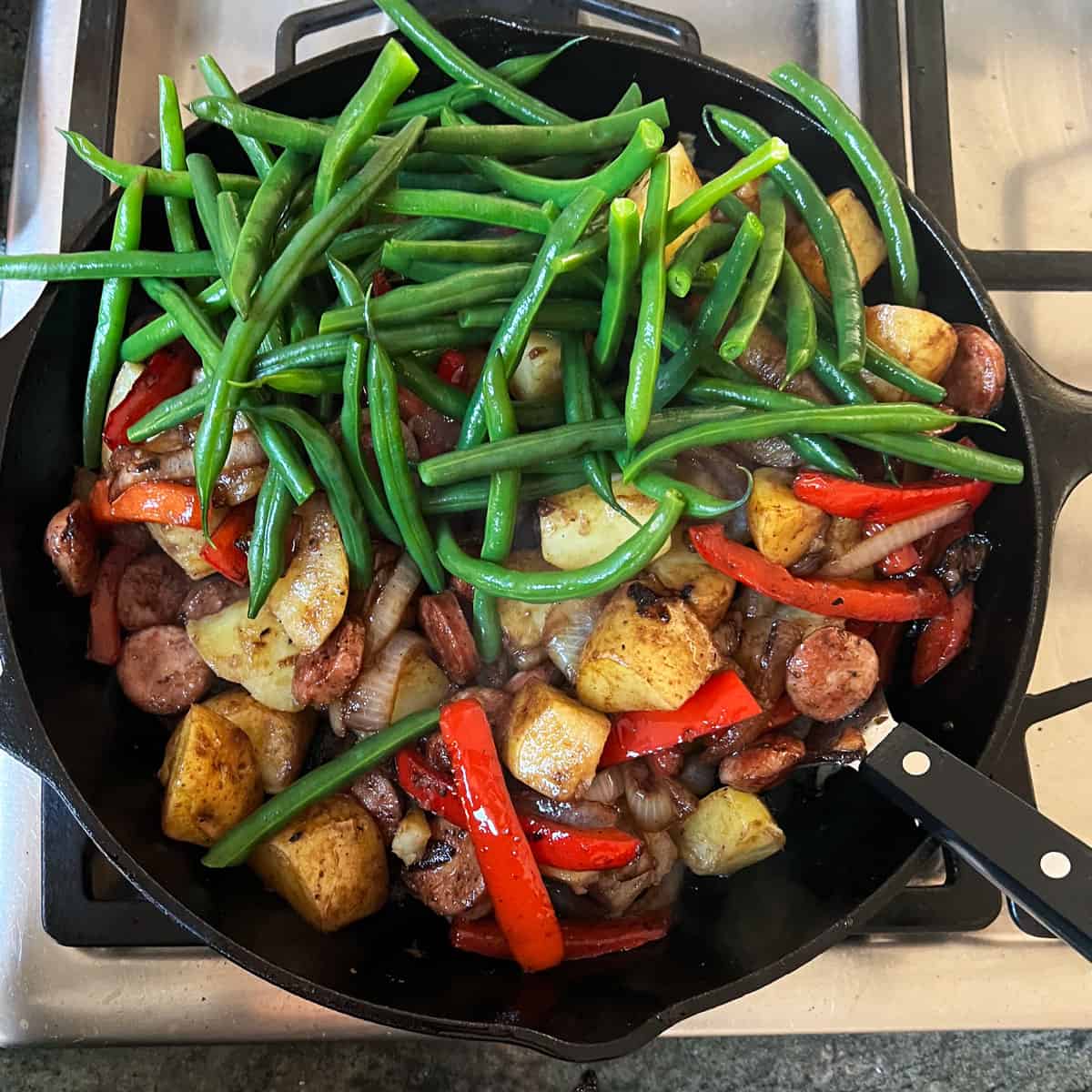 cast iron skillet filled with sautéed sausages, peppers and onions with a pile of blanched green beans on top.