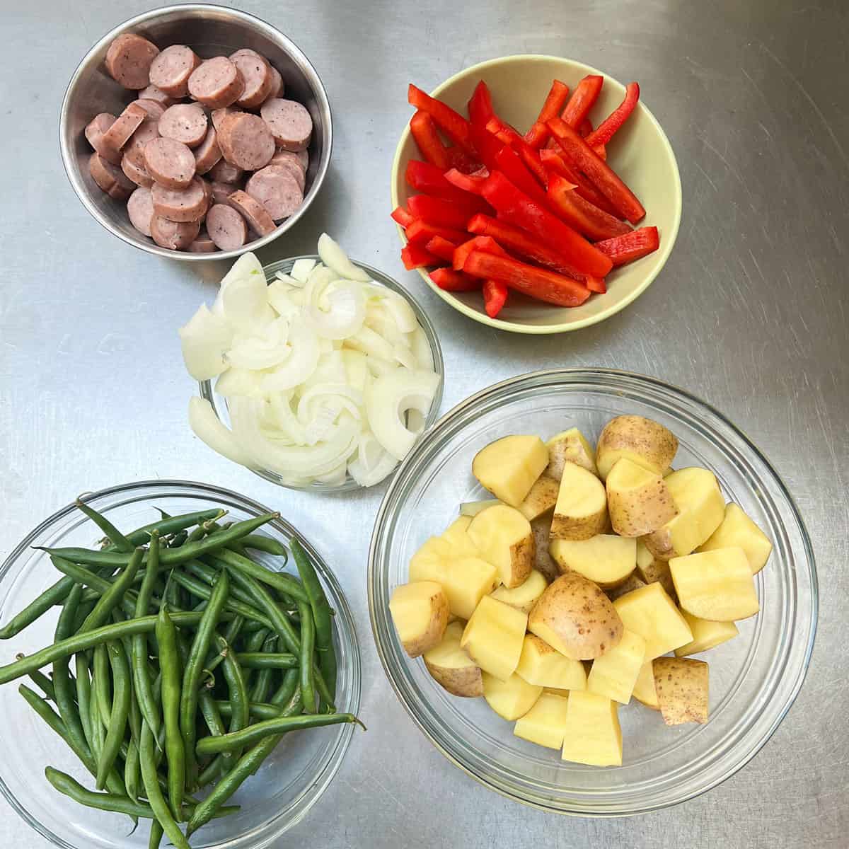 5 bowls on a stainless counter top filled with: red bell pepper strips, sliced onion, cubed gold potatoes, green beans and sliced sausages.