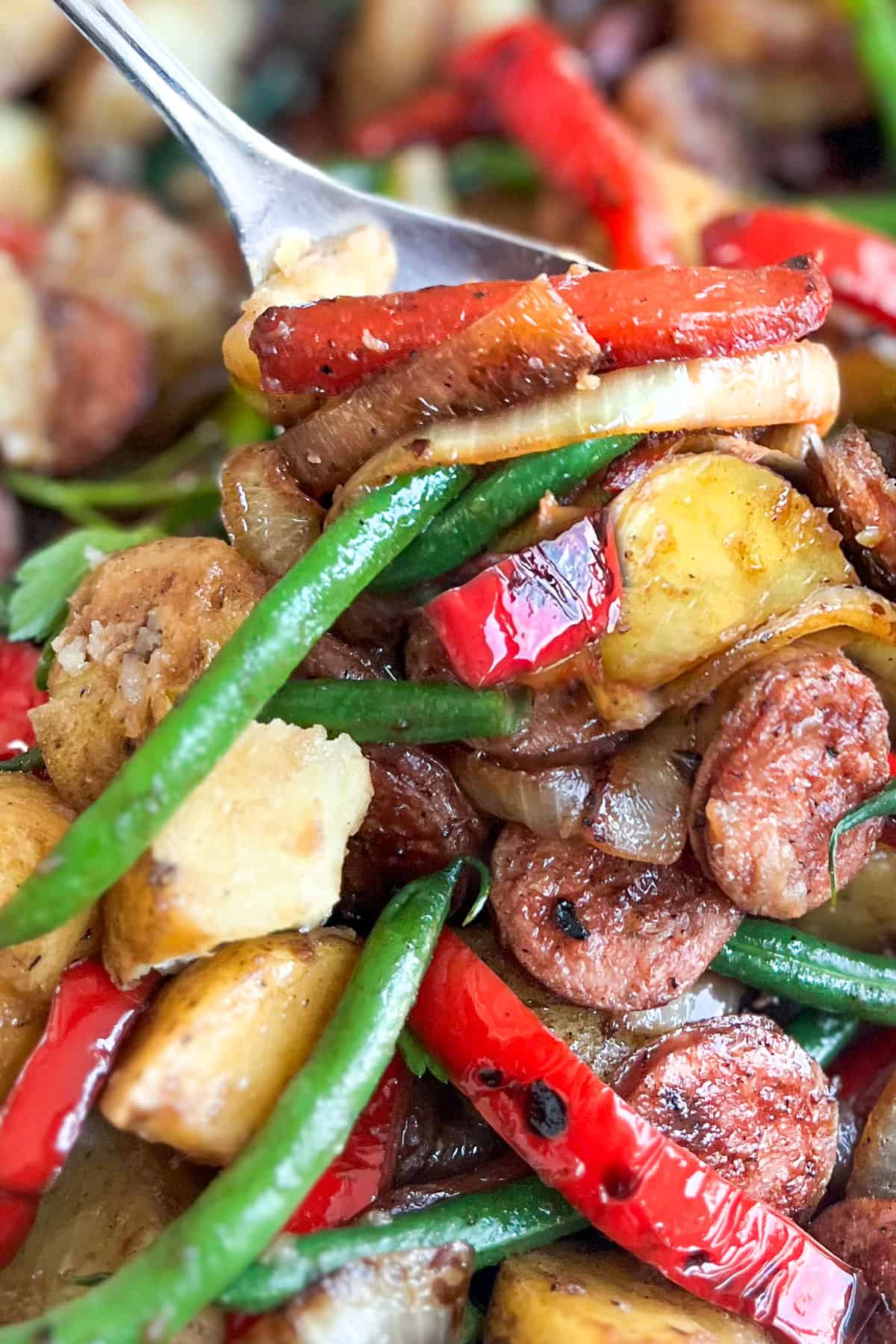 a serving spoon scooping sausages, green beans, potatoes, peppers and onions.