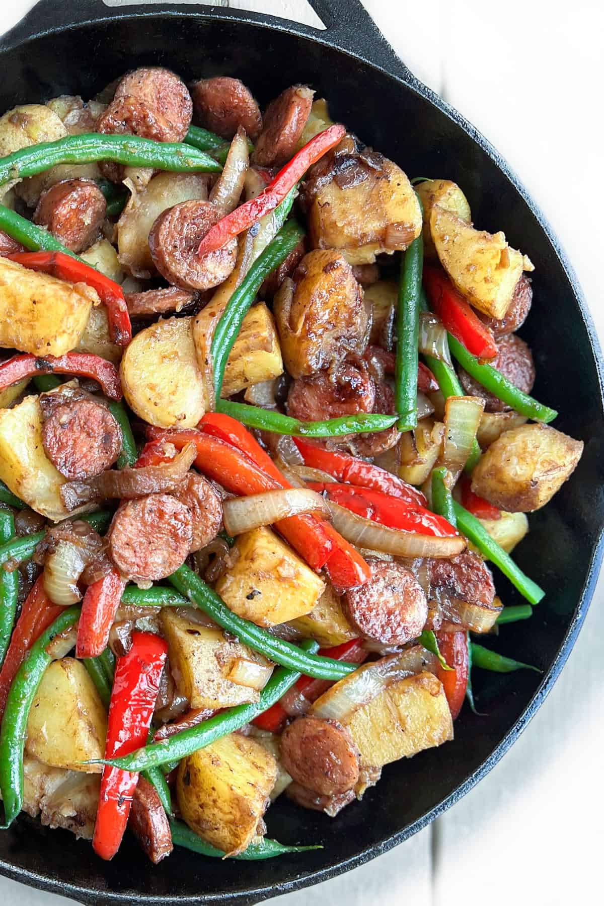 cast iron skillet filled with sautéd slices of sausage, chunks of potato, sliced red bell peppers and green beans