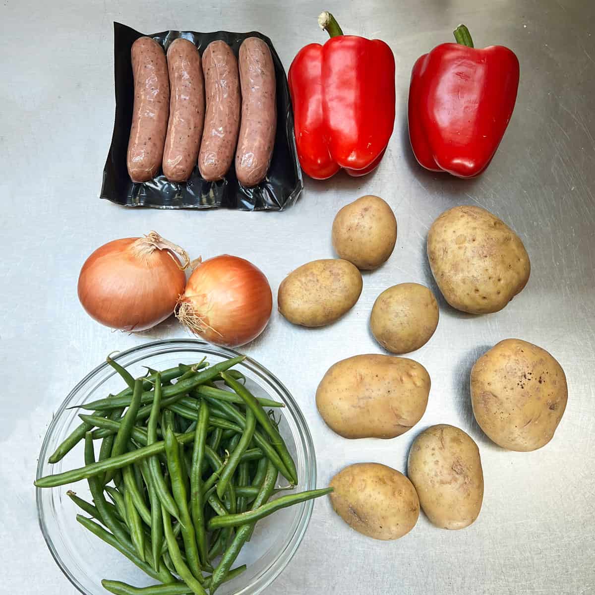 Ingredients on a stainless steel countertop: 4-pack of smoked chicken sausages, 8 yukon gold potatoes, glass bowl filled with green beans, two yellow onions, two red bell peppers.
