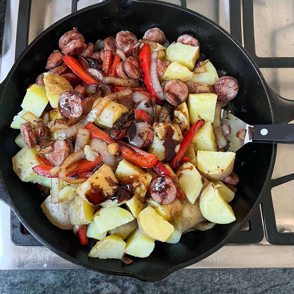 Metal spatula in a cast iron skillet filled with bites sized chunks of boiled potato, strips of red peppers and onions, sauteed sausages and drizzled of pomegranate molasses.