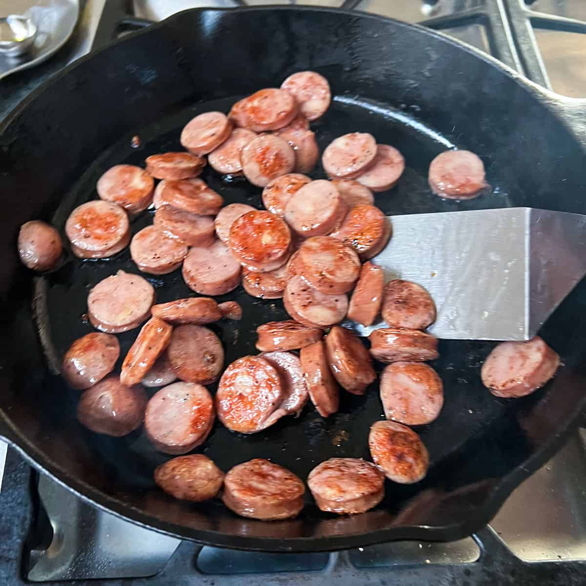 Metal spatula flipping sliced rounds of sauteed smoked sausage in a cast iron skillet.
