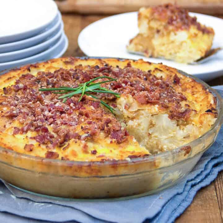 Cauliflower Gruyere pie with bacon parmesan crumb topping in a pie pan with one slice taken out of it and sitting on a plate in the background.