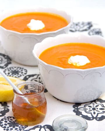 Two bowls of carrot soup topped with a dollop of yogurt, a small jar of honey on the side and half a lemon.
