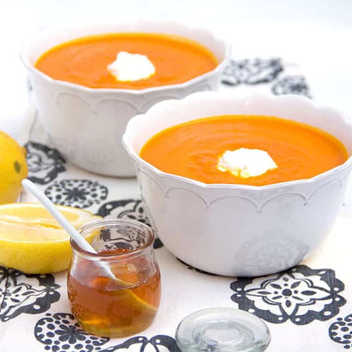 Two bowls of carrot soup topped with a dollop of yogurt, a small jar of honey on the side and half a lemon.