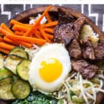 Pinterest pin: photo of a brown wooden bowl filled with colorful bibimbap: carrots, beef, spinach, zucchini, bean sprouts and a fried egg on top