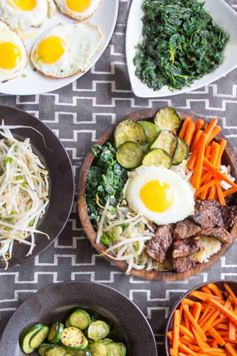 8 bowls of ingredients for bibimbap set out on a grey patterned tablecloth: spinach, bean sprouts, carrots, zucchini, rice, fried eggs, bulgogi beef, and a complete bibimbap bowl