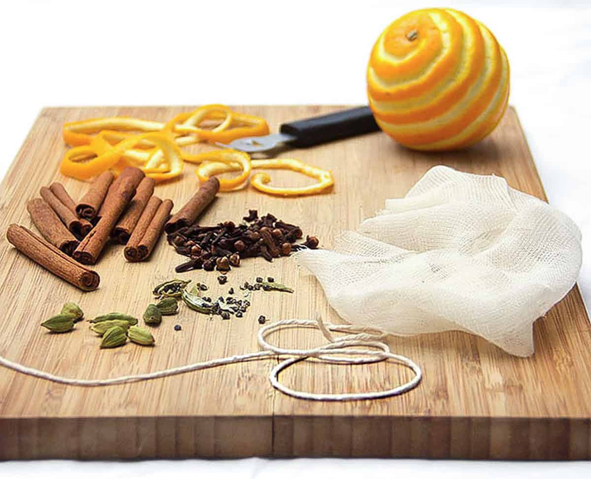 wooden cutting board topped with a piece of cheesecloth, an orange that has been zested. the orange zest, a few cinnamon stick, some cardamom pods, whole cloves and a piece of kitchen twine