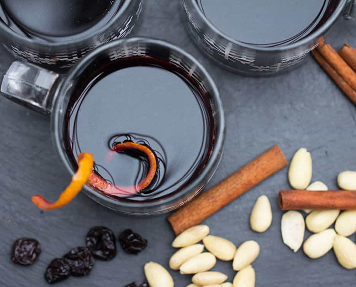 Looking down into a glass of Swedish glogg with a swirl of orange zest, blanched almond, dried cherries and cinnamon stick on the counter around it.