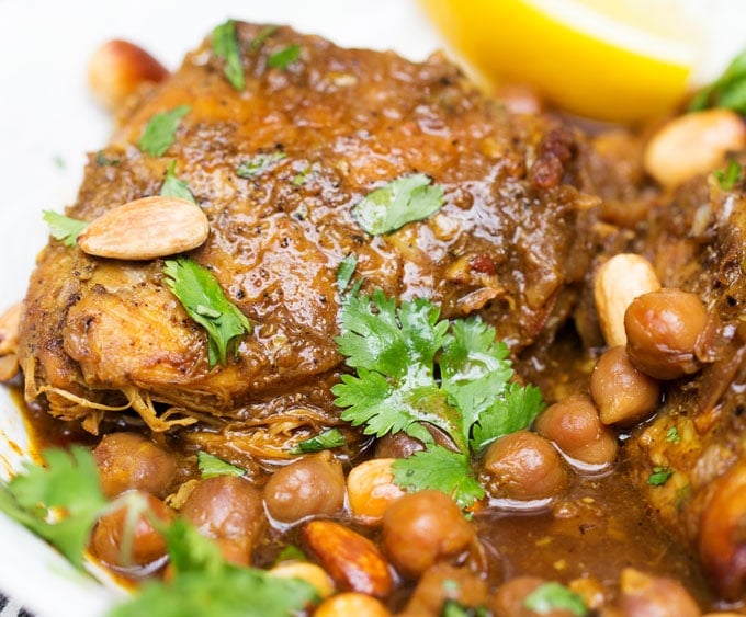 close up of a slow cooked chicken thigh surrounded by chickpeas and rich broth, topped with garnishes of toasted almonds and bits of cilantro and a lemon wedge 