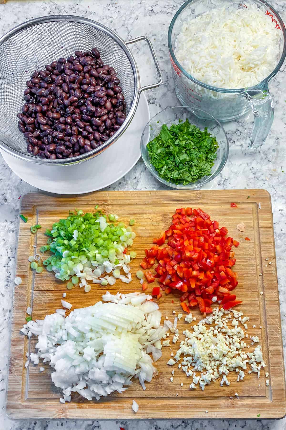 ingredients for gallo pinto chopped and on a wooden cutting board: onions, red pepper, garlic, scallions, a small bowl of chopped cilantro, a large measuring cup of cooked white rice, a colander filled with black beans